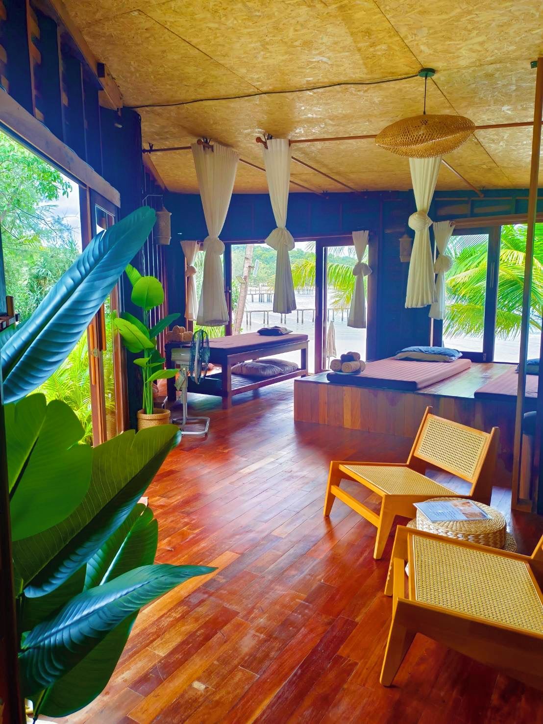 A room with a wooden floor, a bed, a bench, and a dining table. There are large windows and curtains.