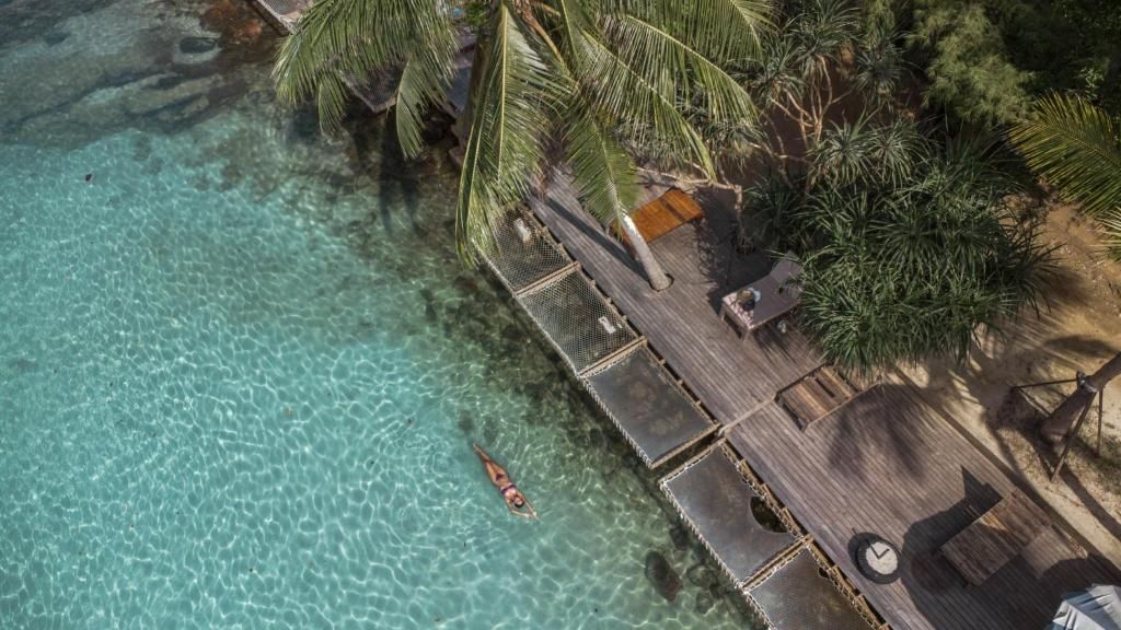 Vue aérienne d’un complexe hôtelier en bord de mer, avec une eau turquoise cristalline, une terrasse en bois, des palmiers et des chaises longues.