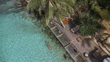 An aerial view of a beach resort with clear turquoise water, a wooden deck, palm trees, and lounge chairs.
