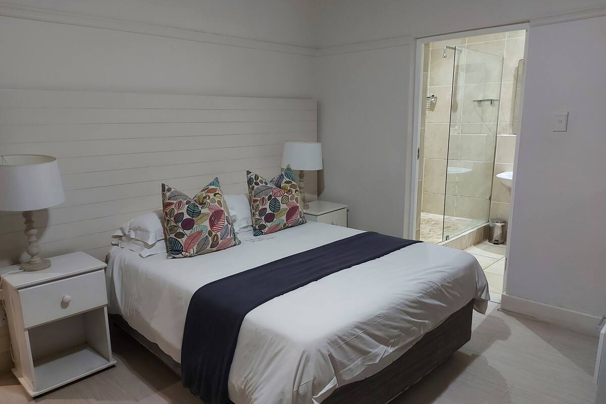 Un dormitorio con cama, dos mesitas de noche con lámparas, una ventana con vistas al baño y una puerta que da a otra habitación.