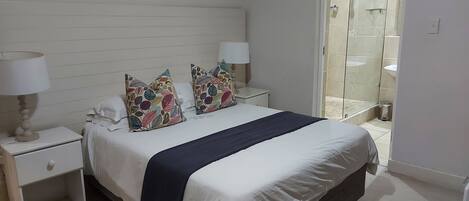 A bedroom with a bed, two bedside tables with lamps, a window with a view of a bathroom, and a door leading to another room.