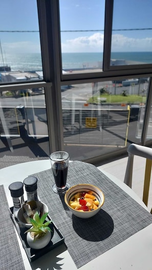 Una mesa con un bol de fruta, un vaso de refresco y condimentos, con vistas al mar y al aeropuerto.