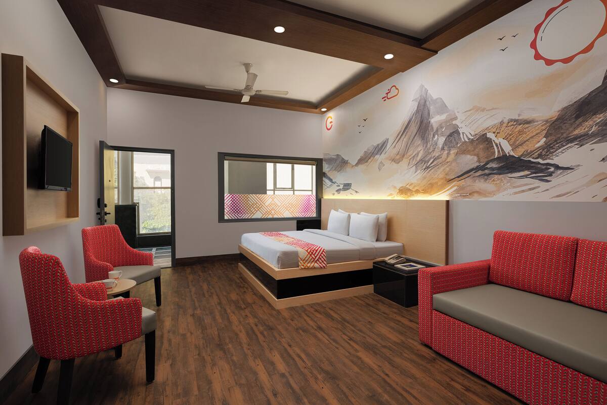 Habitación de hotel con cama, sillones rojos estampados, sofá, televisión y un gran mural en la pared.