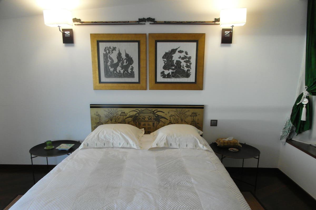 Una cama bien hecha con ropa de cama blanca, dos cuadros en la pared encima y una mesita de noche con una taza verde y un libro al lado de la cama.