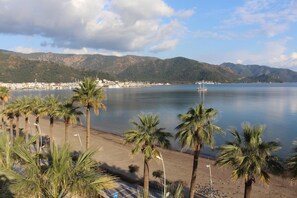 Ein Strand mit Palmen und ruhiger See, eine entfernte Stadt und Berge im Hintergrund.