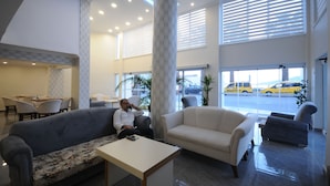 Un hall espacioso con un sofá gris, un sillón color beige y una mesa blanca. Se ven grandes ventanales con persianas, y un área de estacionamiento con taxis amarillos afuera.