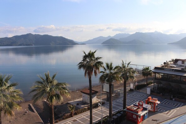 Vista de un lago con montañas al fondo, palmeras en primer plano y un edificio con el letrero "RAMOS" y terraza.