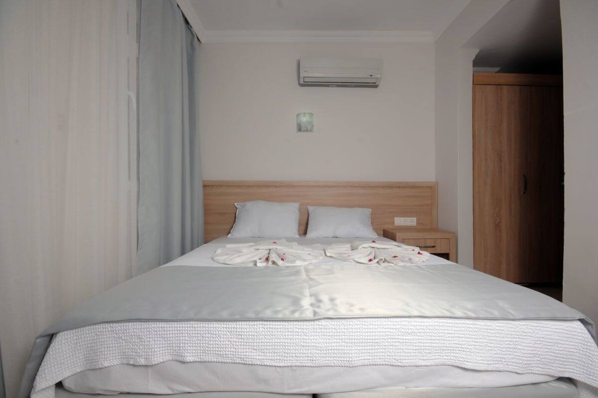 A neatly made bed with white linens, a wooden headboard, a bedside table with a lamp, and an air conditioning unit on the wall.