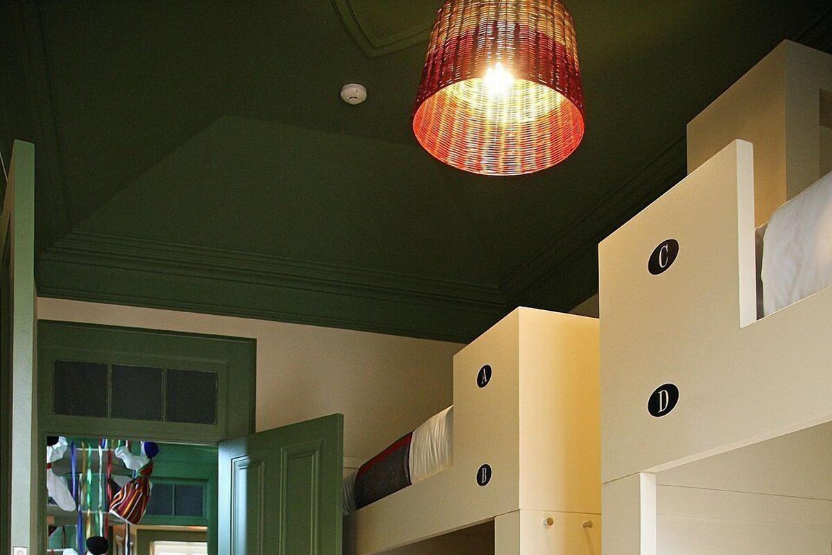 A room with bunk beds, a wooden floor, a green door, and a hanging light fixture.