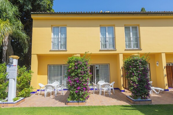 Un edificio amarillo con tres ventanas, sillas blancas y dos macetas con flores moradas.