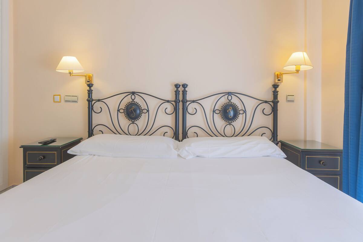 Un lit bien fait, avec une tête de lit en métal, deux tables de chevet avec des lampes et un mur sobre.