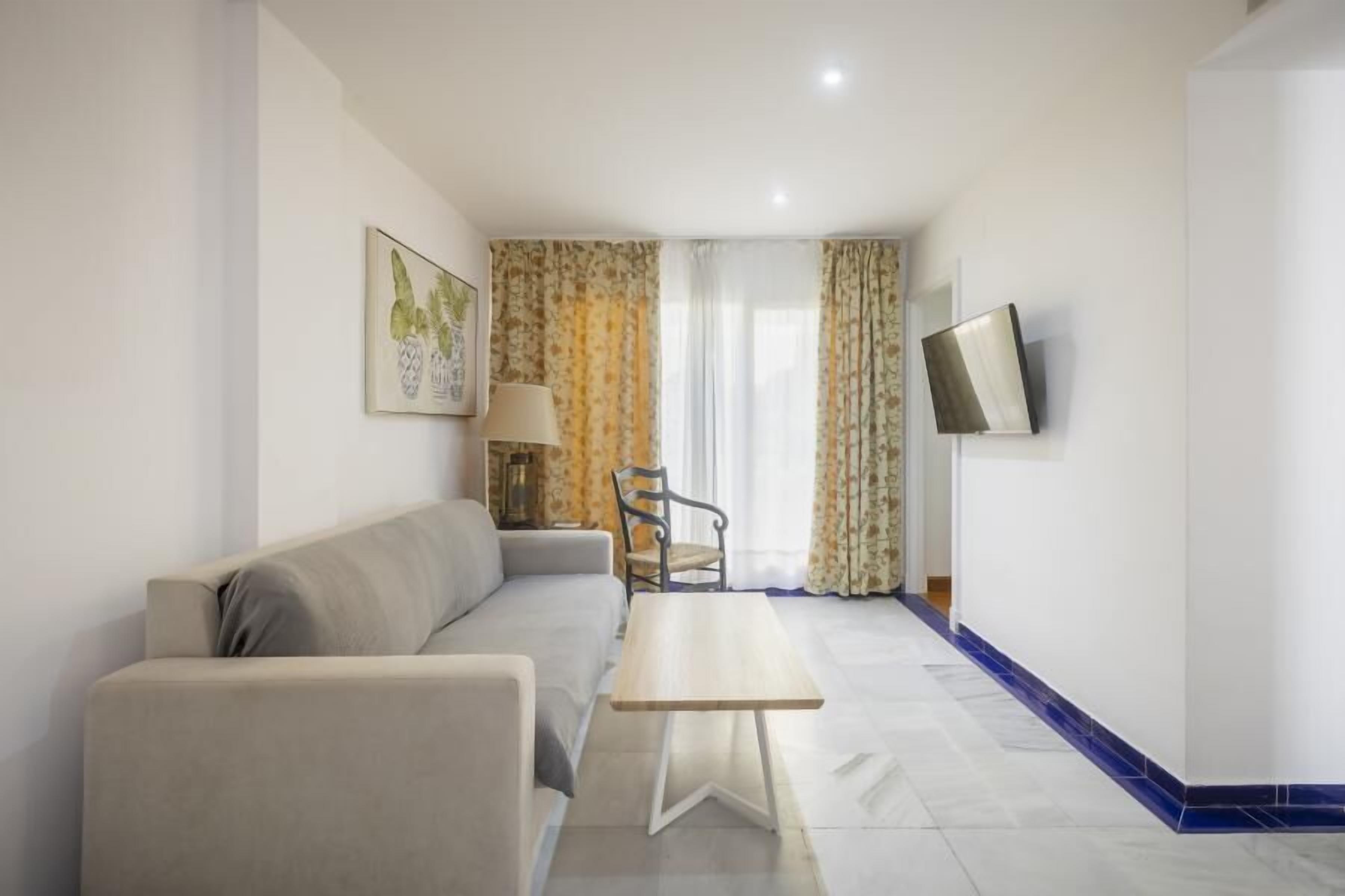 Una sala moderna con un sofá, una mesa de centro de madera, una lámpara y un sillón. Un televisor está montado en la pared.