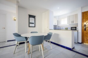 Un comedor con una mesa redonda y cuatro sillas, una cocina con muebles blancos y un microondas, y una puerta de madera.