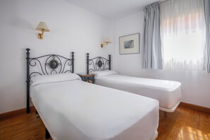 Una habitación de hotel con dos camas individuales, una mesita de noche pequeña con lámpara, un cuadro en la pared y una ventana con cortinas.