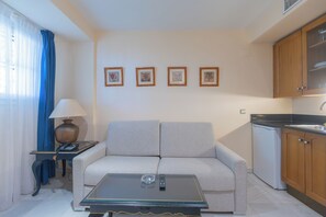 Una sala de estar con un sofá, una mesa de centro, una lámpara y una zona de cocina con armarios y un refrigerador.