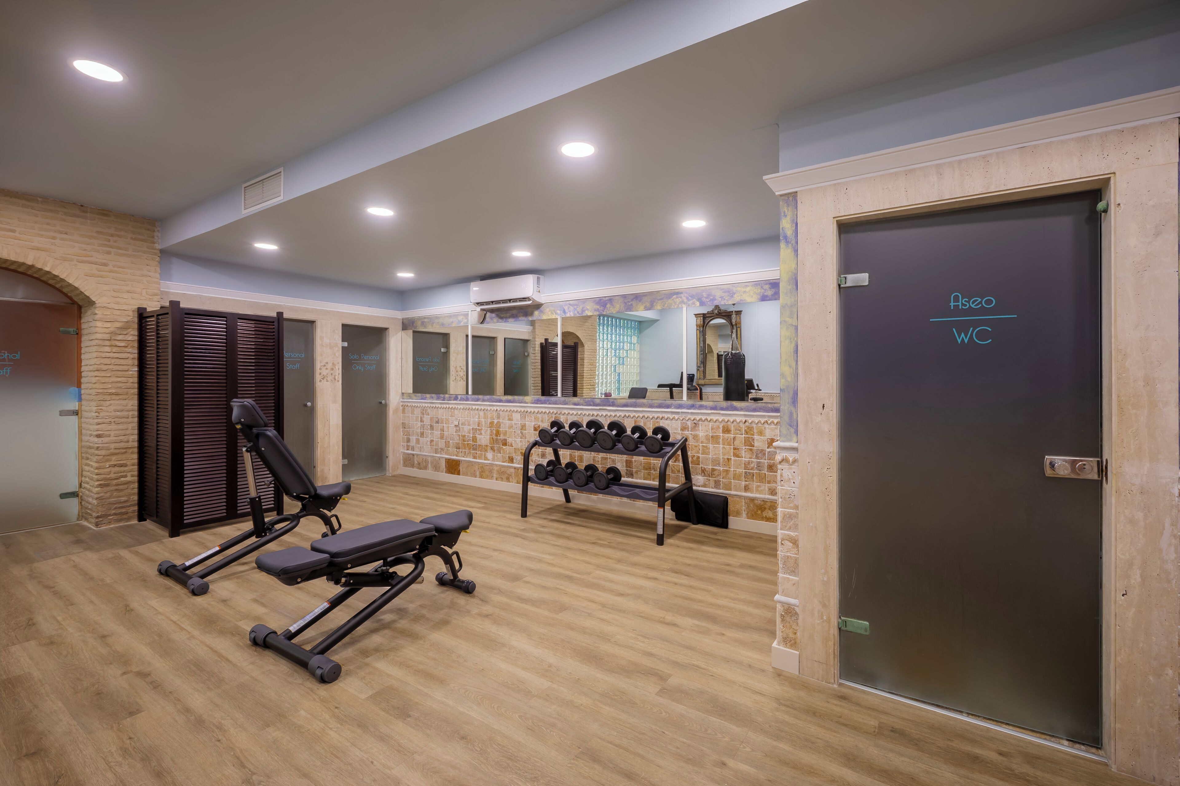 Un gimnasio con equipo de ejercicio, una banca, un estante con mancuernas y una puerta que indica 'Aseo WC'.