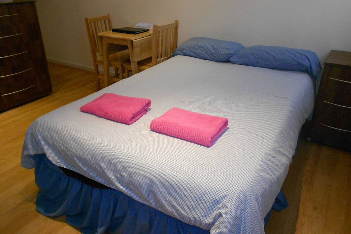A neatly made bed with a blue bedspread and two pink towels, a wooden chair, and a small table with a book and a tissue box.