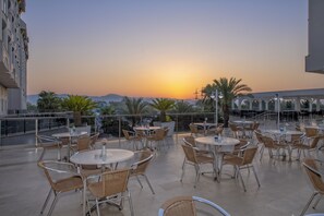 Una terraza al aire libre con mesas redondas y sillas de mimbre, con vistas a una puesta de sol y montañas distantes.