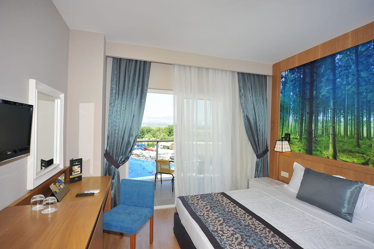 A hotel room with a bed, a desk with a TV, a chair, a balcony with a view of a swimming pool, and a large forest-themed wall mural.