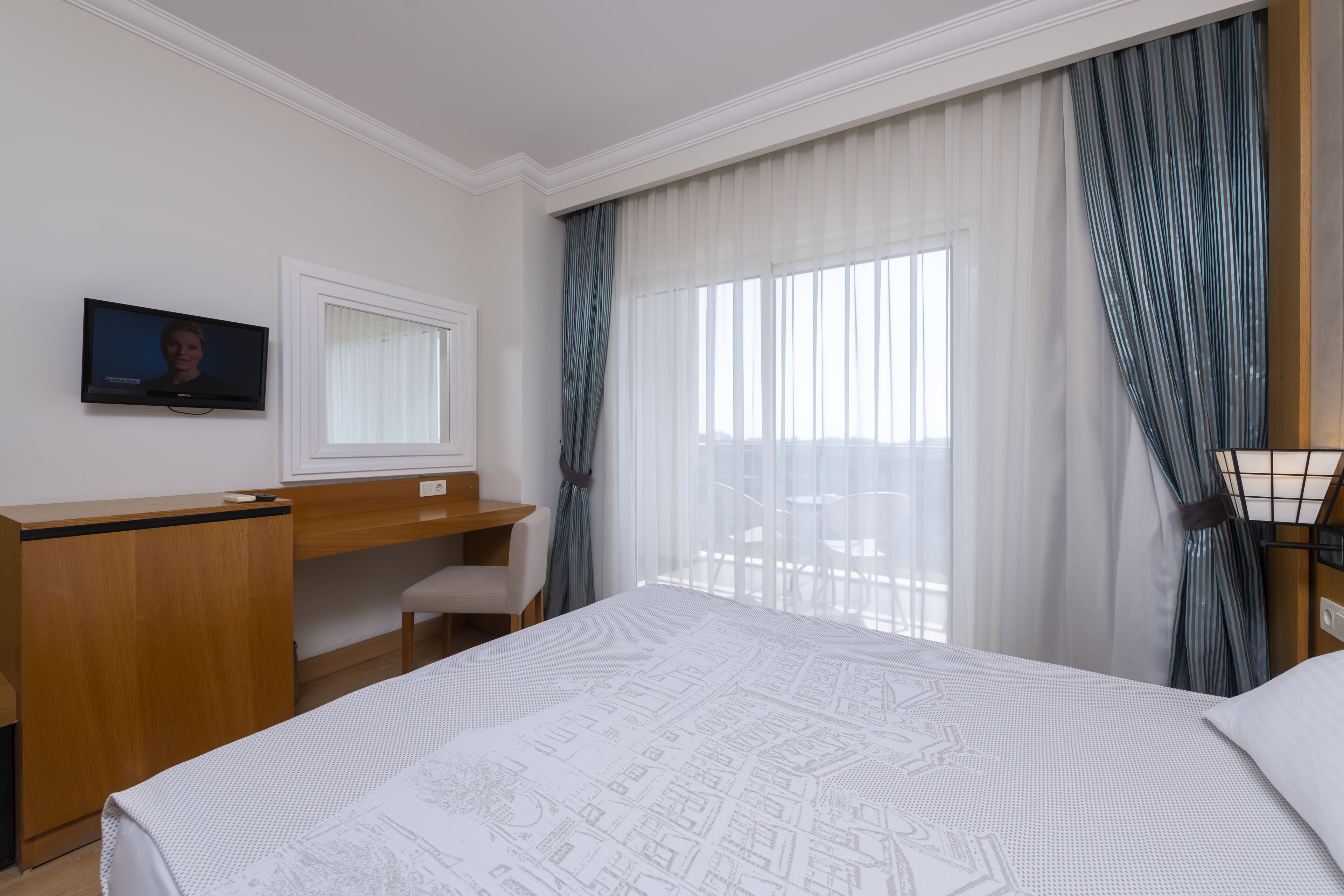 Ein Hotelzimmer mit einem Bett, einem Schreibtisch, einem Sessel, einem Fernseher und einem Vorhangfenster.