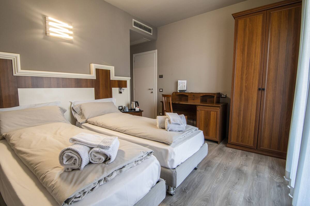 A hotel room with two beds, a wooden wardrobe, a desk, and a nightstand.