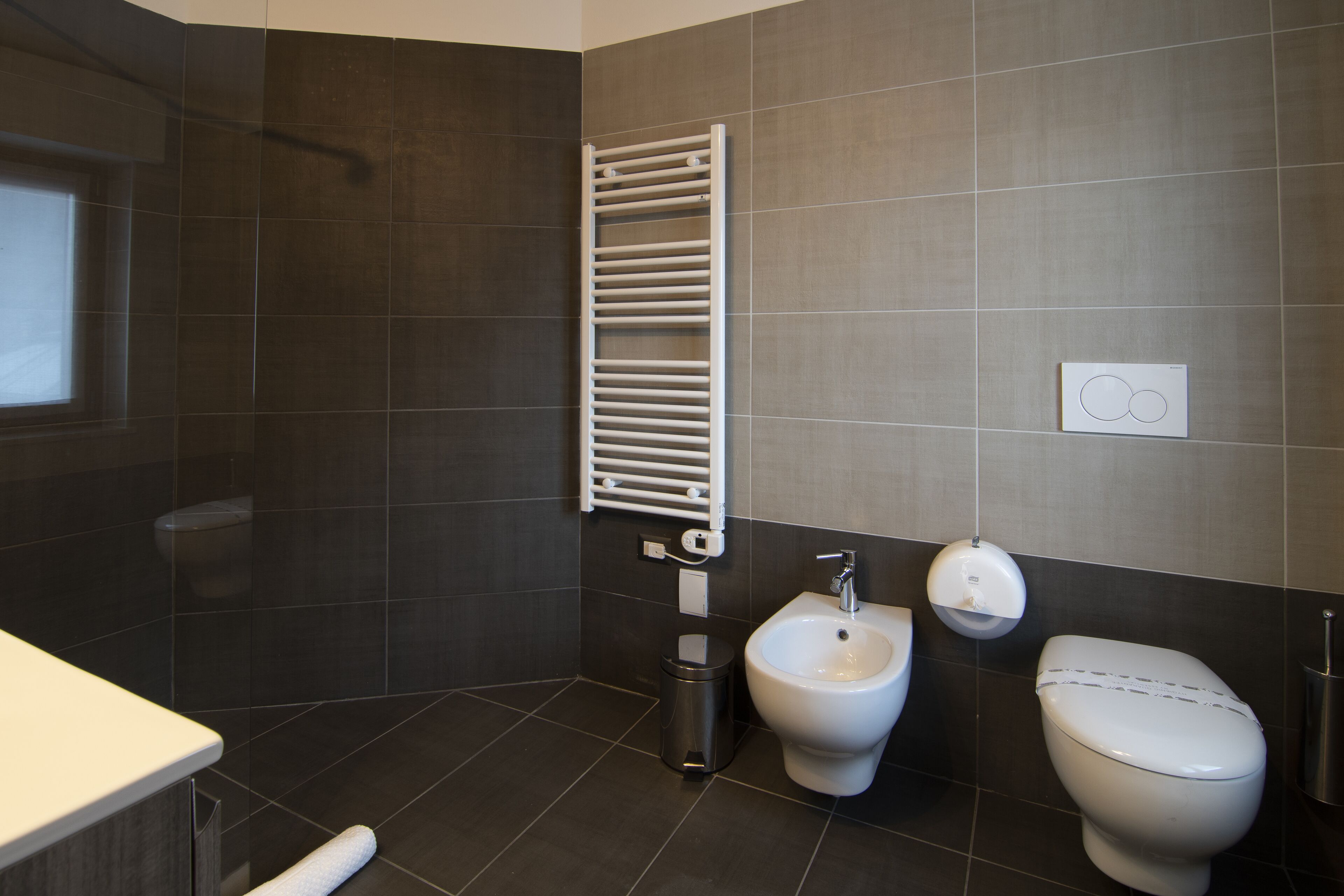 Un baño moderno con baldosas oscuras, un tendedero blanco, un bidé, un inodoro y un cubo de basura.