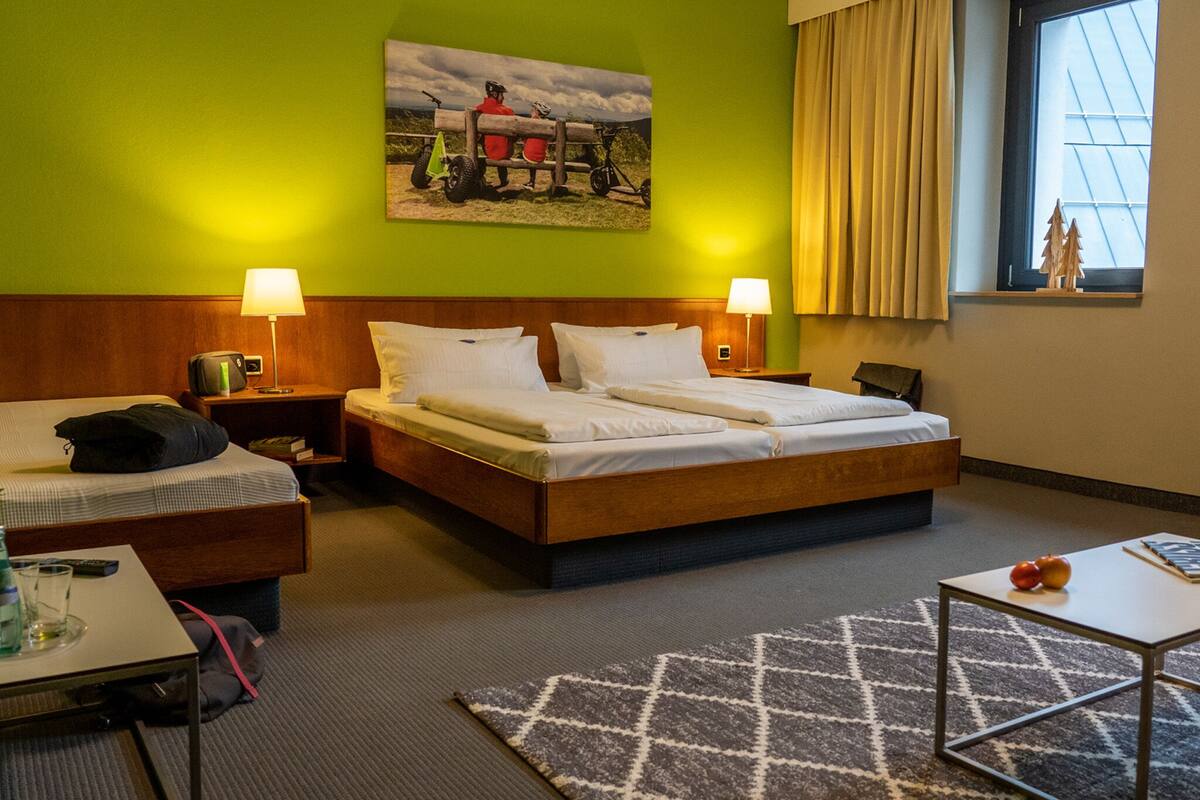 A hotel room with a bed, two bedside tables, a nightstand, a desk, a chair, a window with curtains, and a wall-mounted picture.