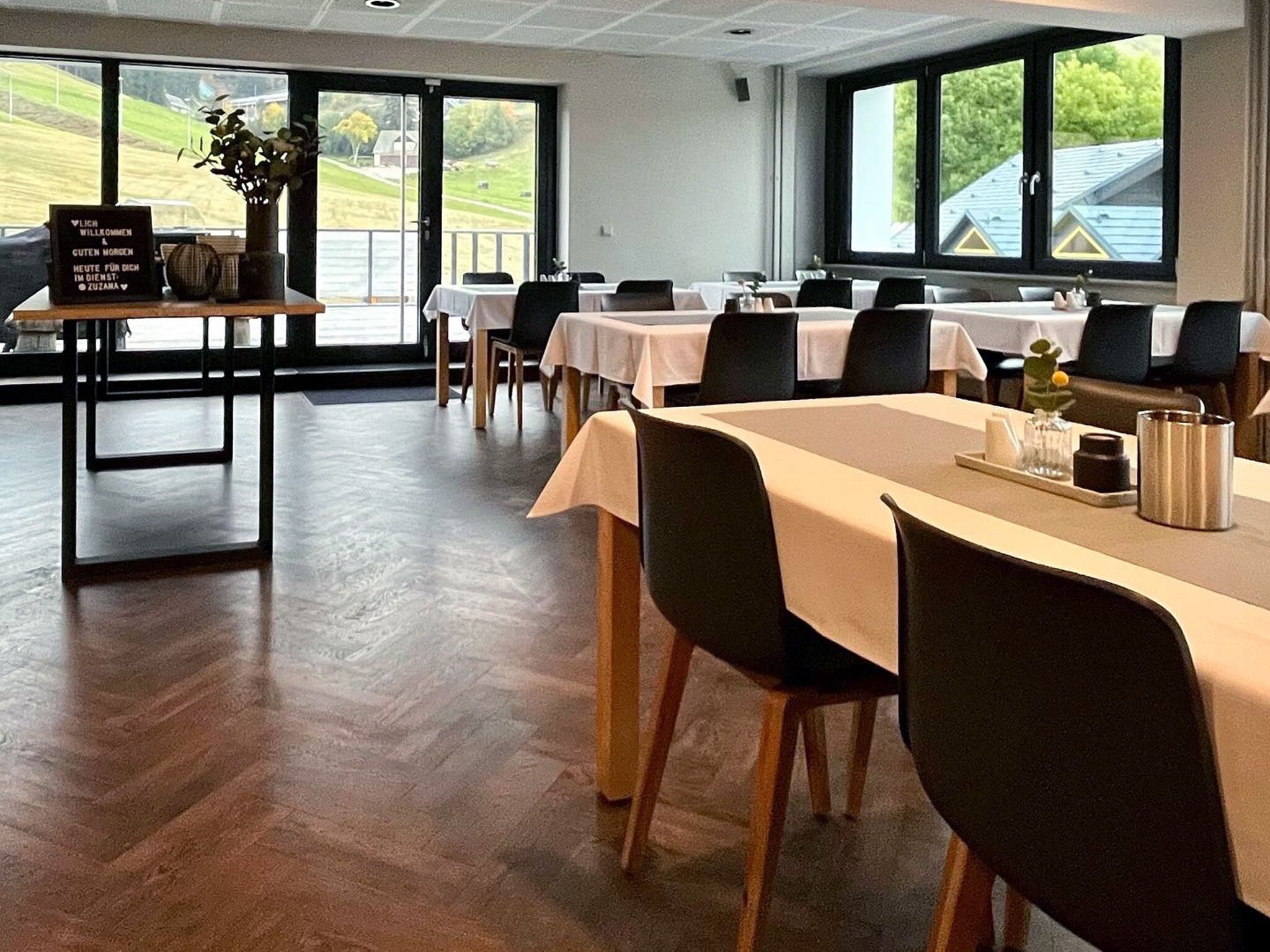 A dining area with tables set for guests, large windows with a view of the outdoors, and a sign on the table.