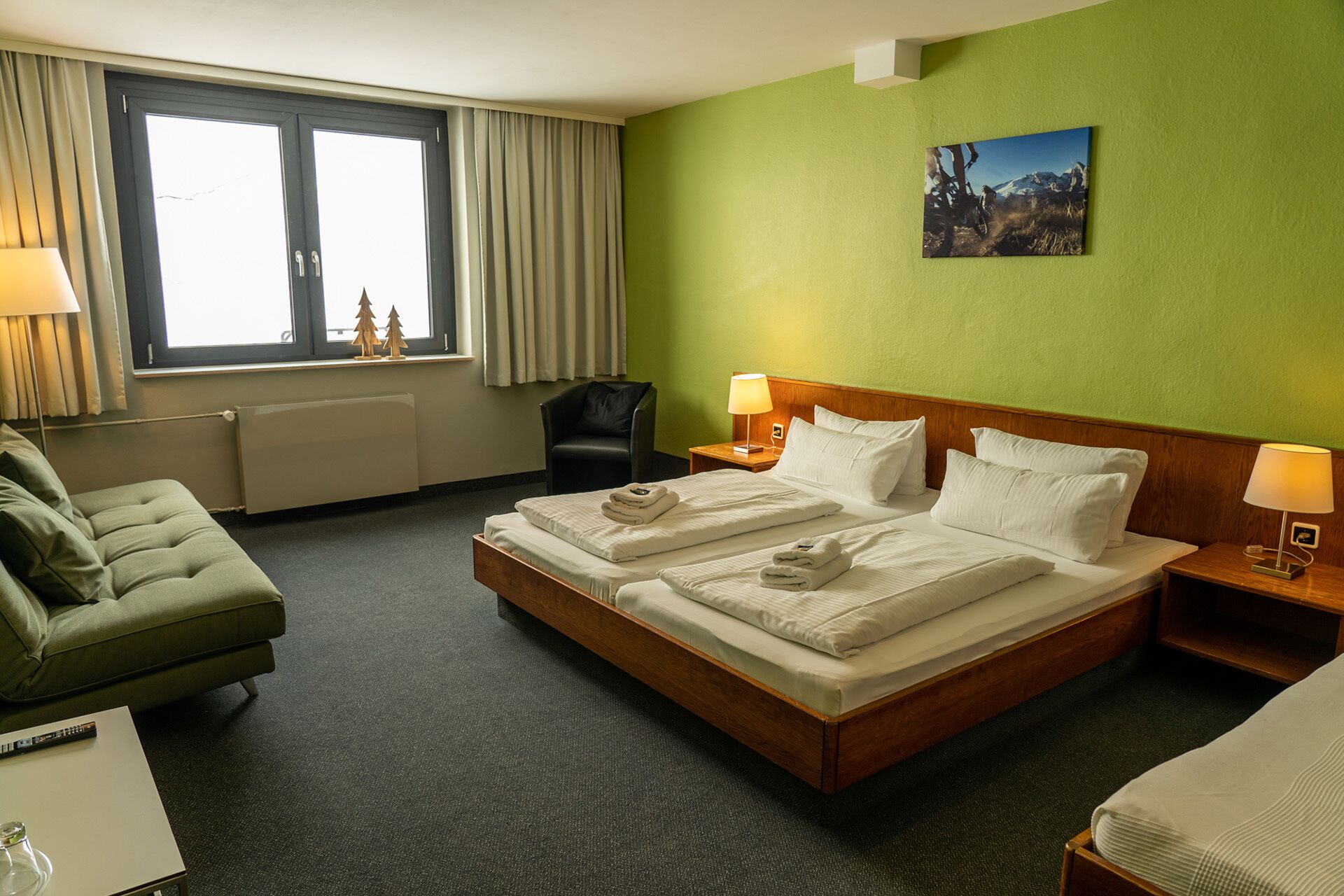 A hotel room with a large bed, two bedside tables with lamps, a green sofa, a window with curtains, and a picture on the wall.