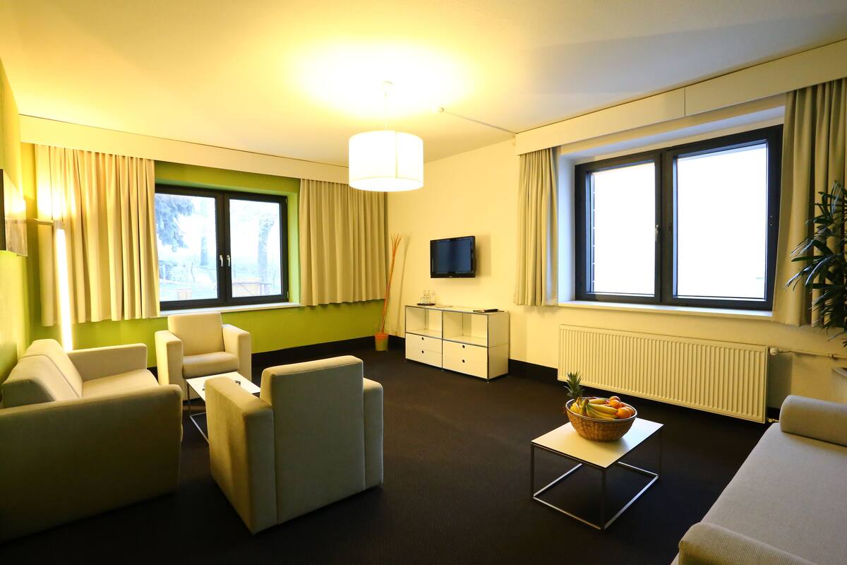 A modern hotel room with a sofa, armchairs, a small table with a fruit bowl, a TV, and large windows with curtains.