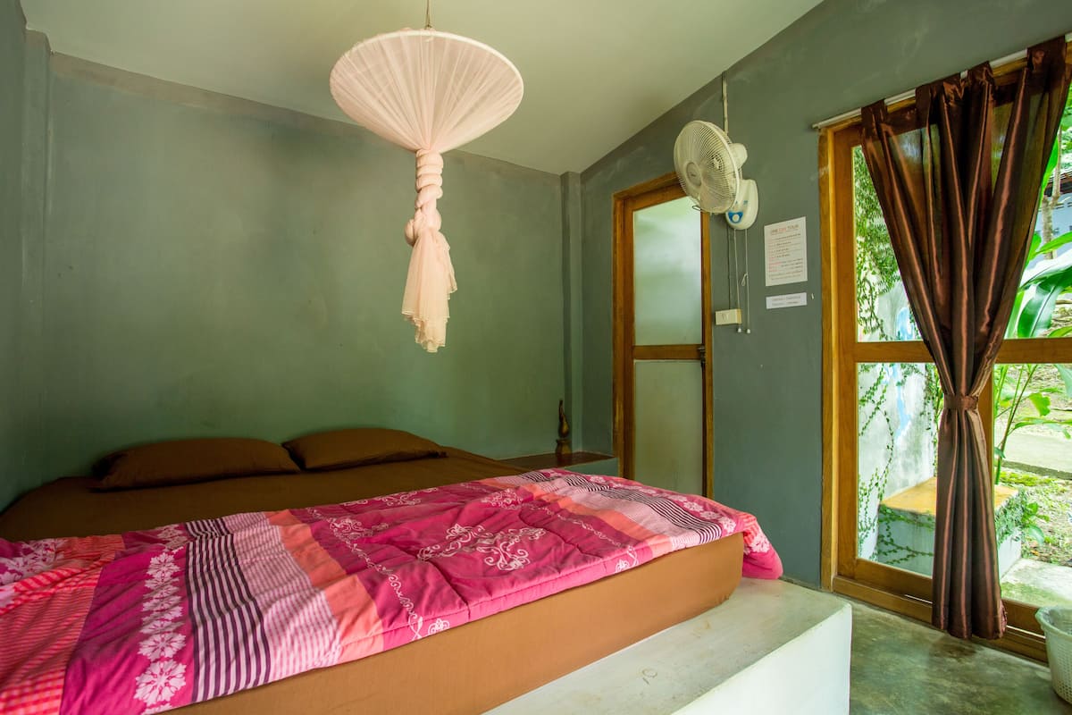 A bedroom with a bed, a hanging light fixture, a window with curtains, and a fan on the wall.