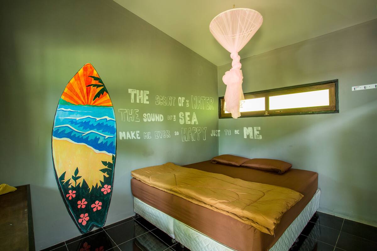 A bedroom with a bed, a wall with inspirational text, a surfboard decoration, and a hanging light fixture.