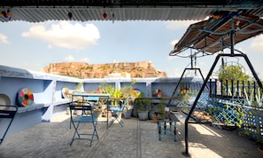 A rooftop terrace with a view of a historic fort, blue chairs, potted plants, and a swing set.