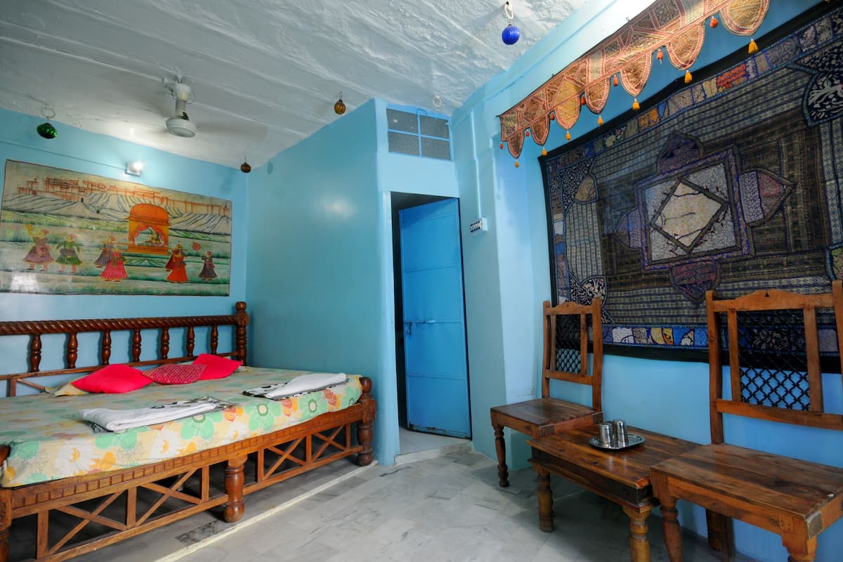 A room with a bed, a wooden table, a chair, a blue door, and a wall with a detailed tapestry.
