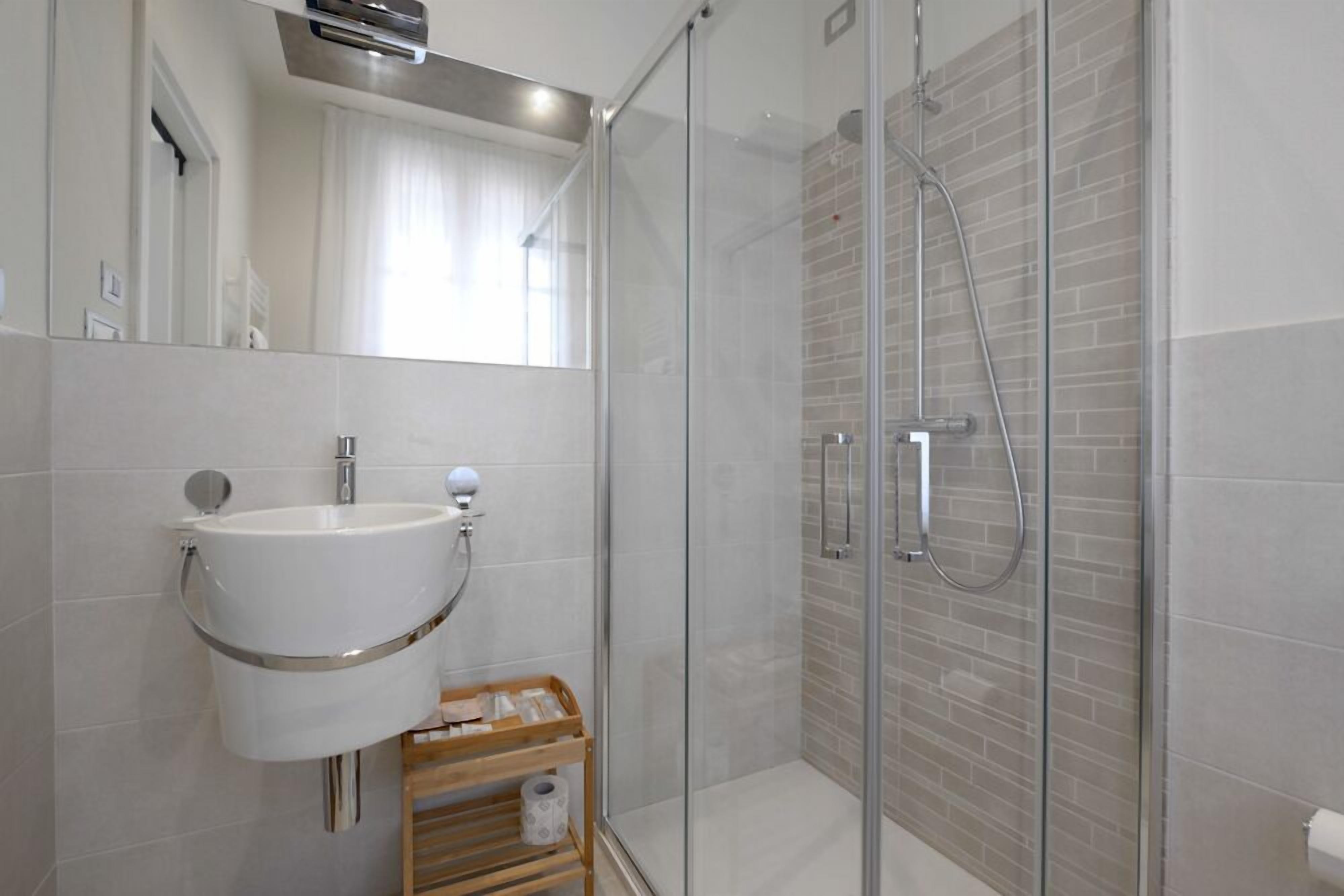 Un baño moderno con ducha de vidrio, lavamanos blanco con grifería cromada y un pequeño estante de madera con artículos de aseo.