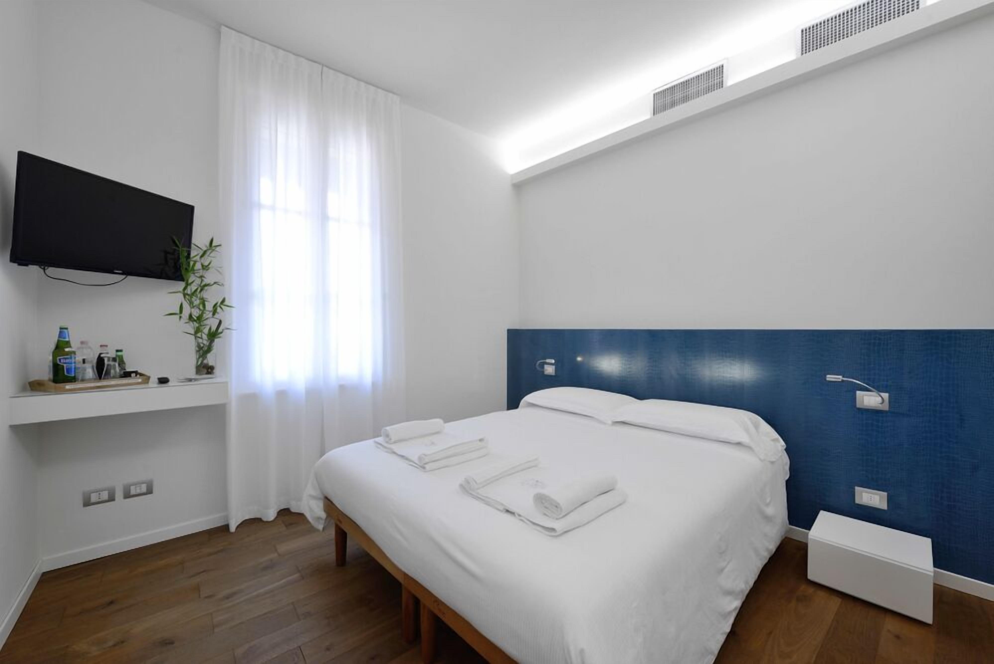 Ein Hotelzimmer mit Bett, Wandfernseher, Fenster mit Vorhängen, Ablagefläche mit Toilettenartikeln und blauer Akzentwand.