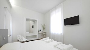 A modern hotel room with a large bed, a flat-screen TV mounted on the wall, a small seating area with a chair, and a bathroom visible through an open door. - La corte sul naviglio (Boffalora Sopra Ticino)