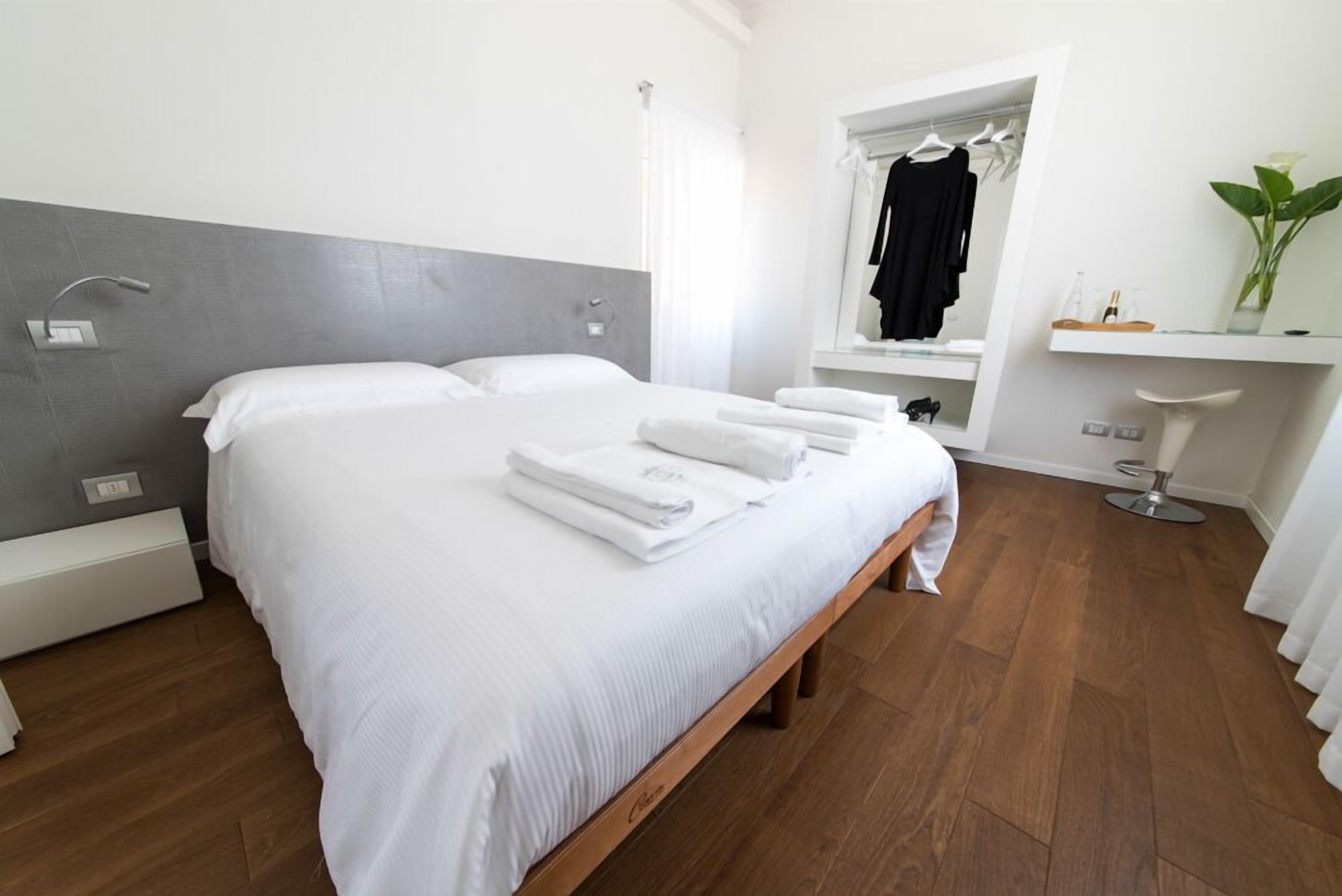 Una habitación de hotel moderna con una cama grande, una mesita de noche, un espejo sobre la cama, una zona de baño pequeña y una planta en un estante.