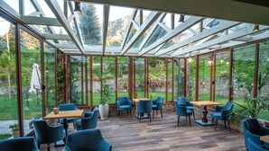 A conservatory with wooden flooring, blue chairs, and tables, featuring a glass ceiling and a view of a garden and mountains. - Anavasi Mountain Resort (North Tzoumerka)