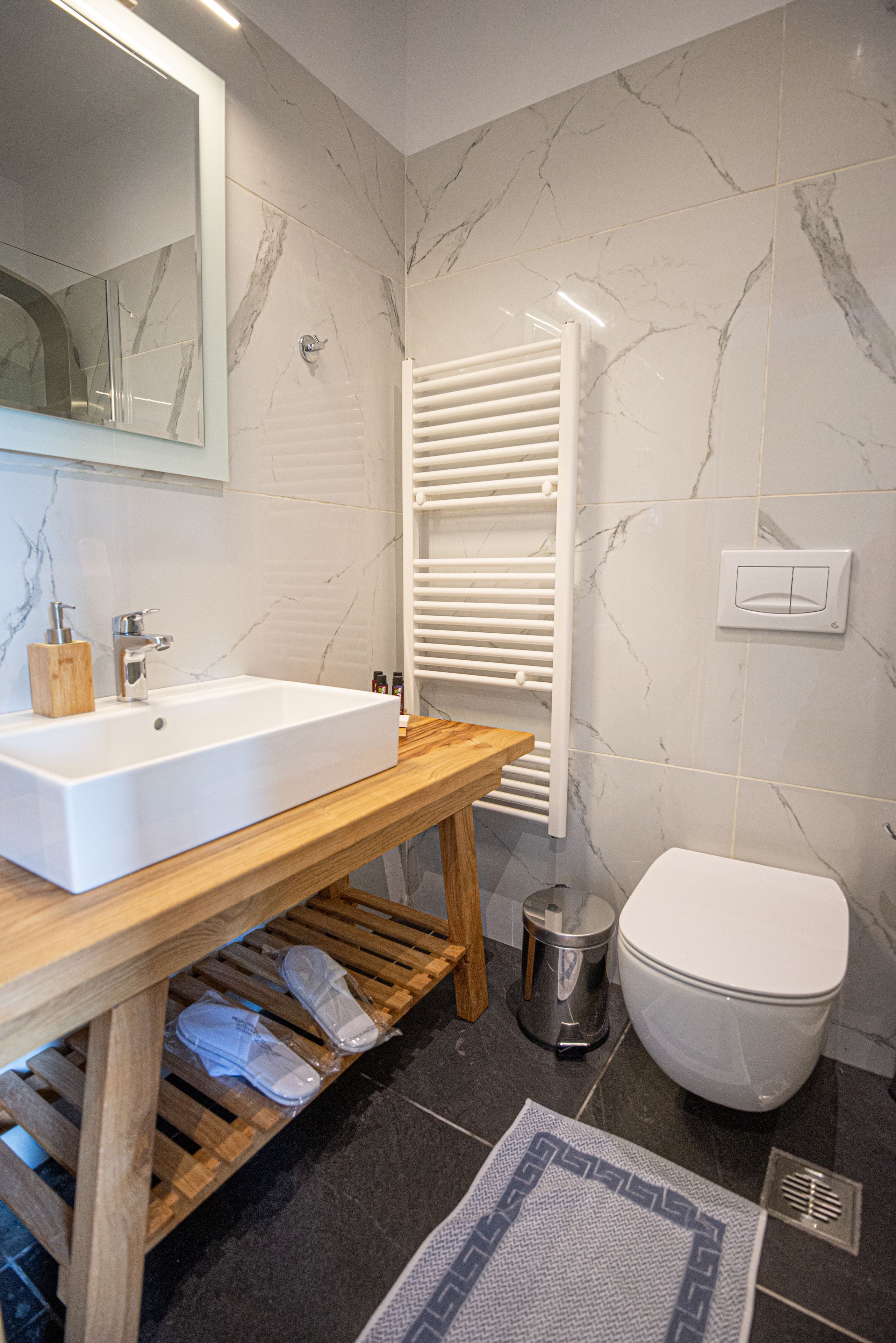 Un baño moderno con lavamanos blanco, tocador de madera, paredes de mármol, inodoro blanco y un perchero para toallas.
