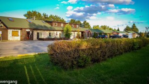 A row of rustic cabins with stone walls and green roofs, a parking area, and a clear sky.