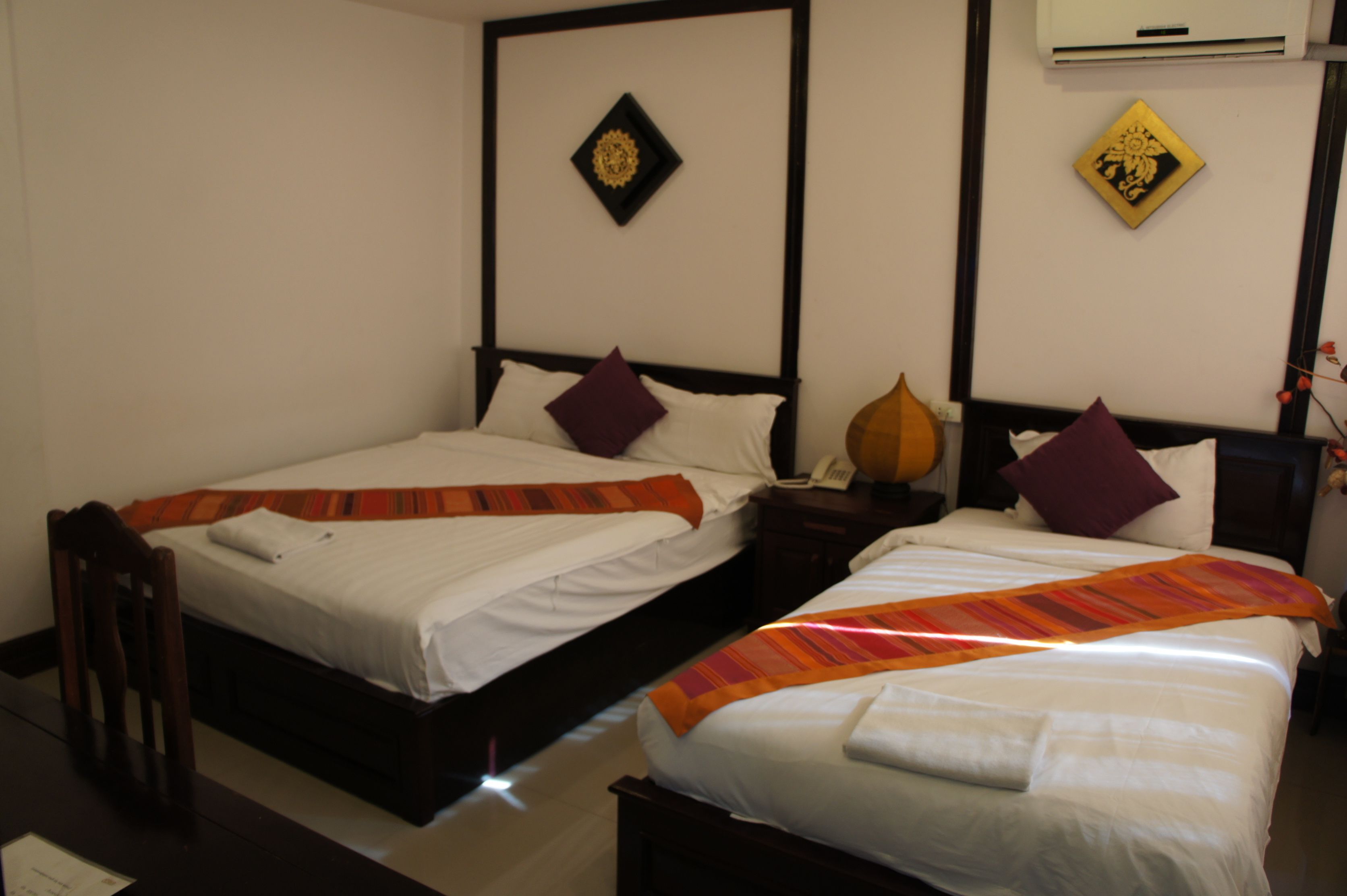 Two single beds with wooden frames, white bedding, and decorative pillows. There is a wall-mounted air conditioner and two wall decorations.