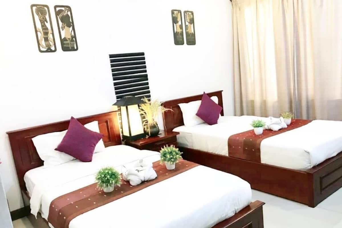 Quarto de hotel com duas camas, cada uma com cabeceira de madeira, roupa de cama branca e manta marrom. Há duas mesas de cabeceira com luminárias e objetos decorativos.