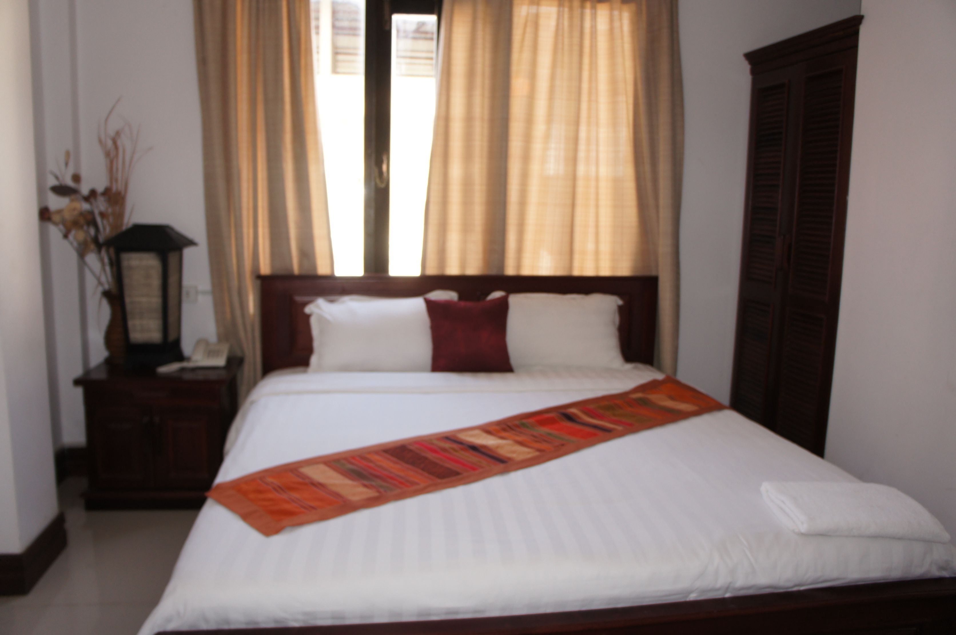 A neatly made bed with a white comforter and a decorative throw, a wooden nightstand with a lamp, and a window with beige curtains.
