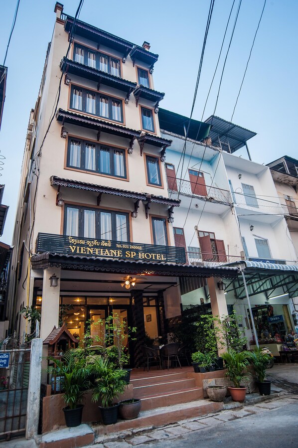 A multi-story building with a sign for "VIENTIANE SP HOTEL" at the ground level. There are steps leading to the entrance, and potted plants are placed in front.