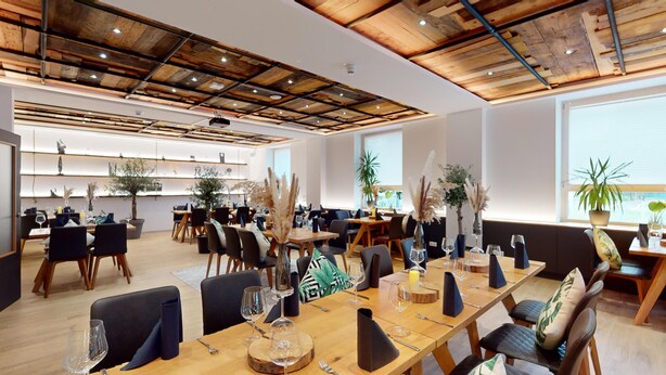 Een modern restaurant met houten tafels en stoelen, potplanten en een hoge plafond met zichtbare balken.