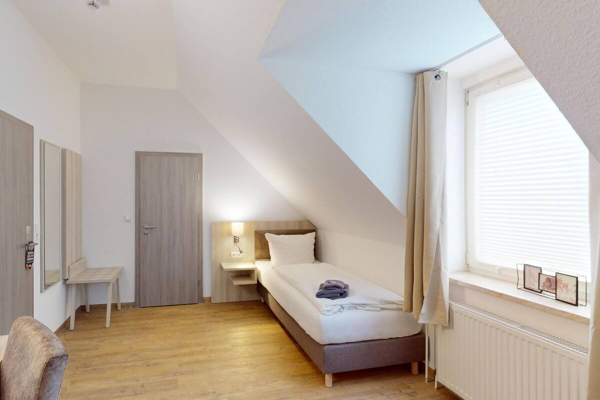 Una habitación pequeña y bien iluminada con una cama, un escritorio y una silla. Cuenta con una ventana con persianas y un radiador debajo.