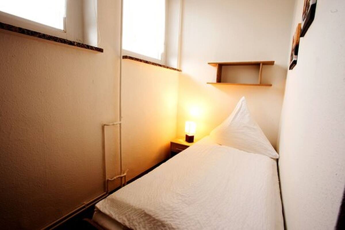 Una habitación de hotel pequeña y acogedora con una cama individual, una mesita de noche con una lámpara y un estante pequeño sobre la cama.