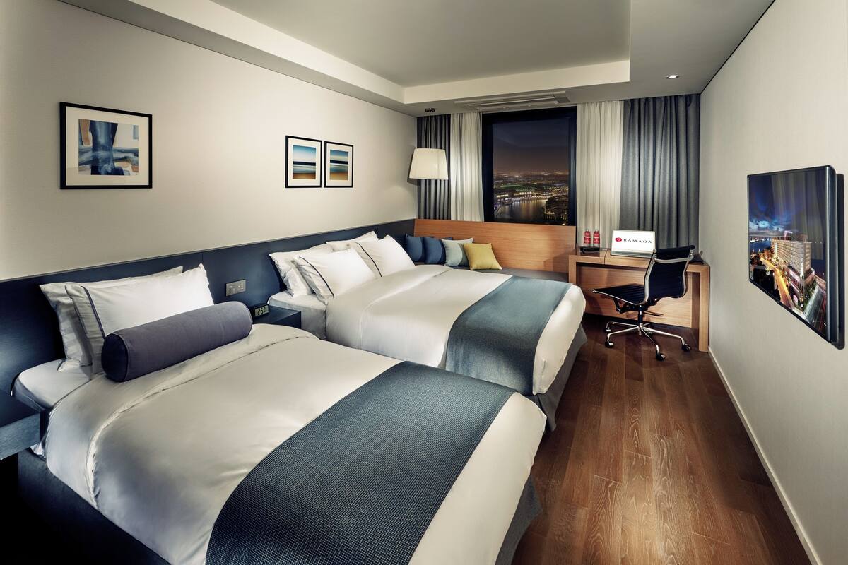 A hotel room with two beds, a desk, a chair, a lamp, and a city view.