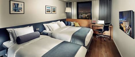 A hotel room with two beds, a desk, a chair, a lamp, and a city view.